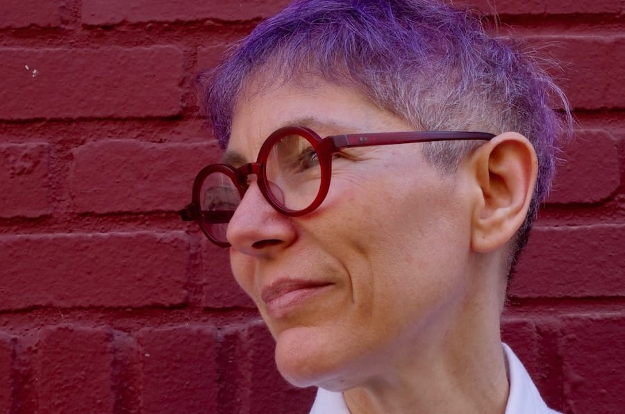 headshot & shoulders of Emma head turned away in a white button-down shirt with red glasses and short purple hair against a red brick wall