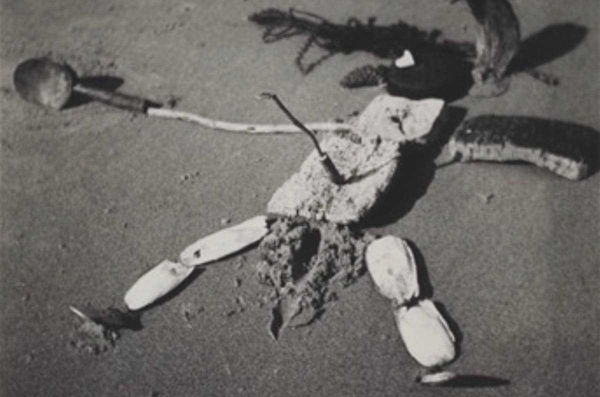 Vintage photo of a crude sculpture from beach detritus, of a figure lying on its back, and pierced with a stick in its belly.