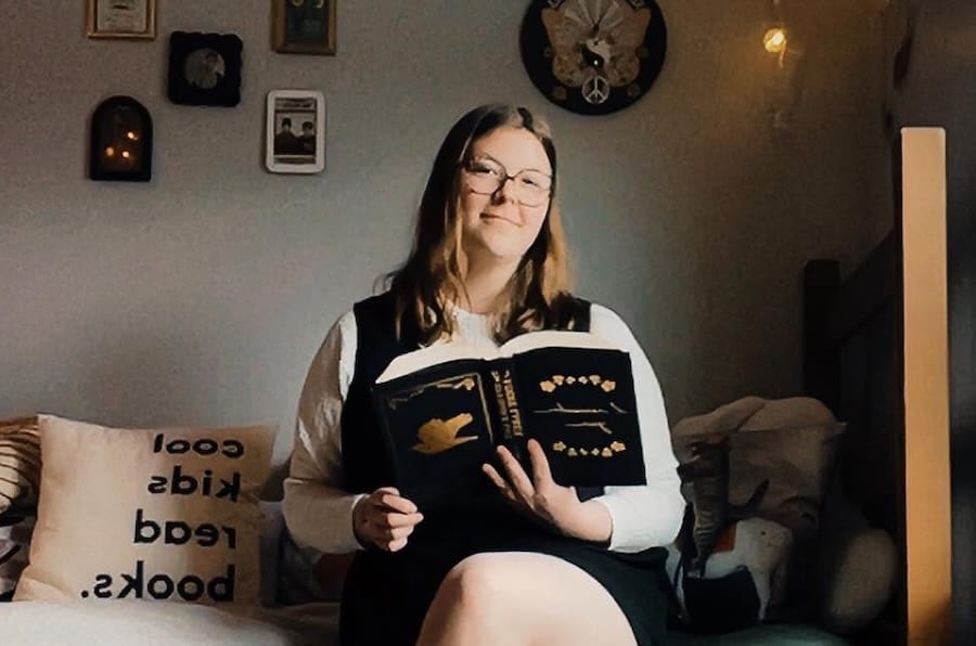 Courtney sits on a bed with a book open in hand, looking and smiling at the camera.